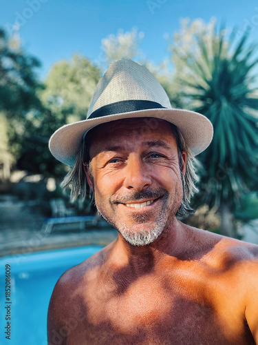 Happy blond man with straw hat and a stubble beard by a pool in a sunny summer garden.