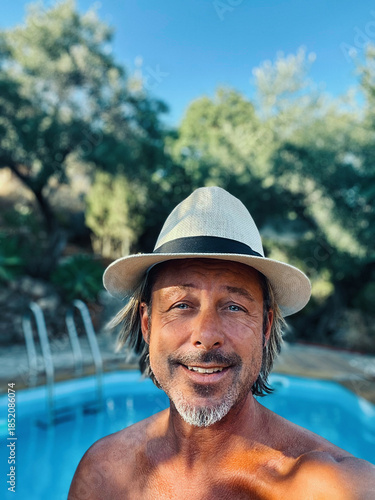 Happy blond man with straw hat and a stubble beard by a pool in a sunny summer garden.