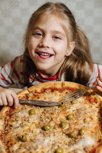 Happy little girl eating homemade pizza with cheese and olives at the table