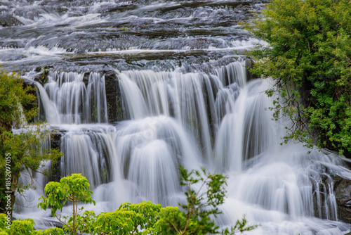 Silky smooth waterfall cascading over rocky ledges with long exposure motion blur effect surrounded by vibrant green foliage and lush vegetation in pristine natural wilderness stream river landscape