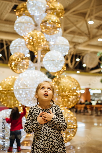 Little girl amazed by glowing Christmas lights in shopping mall, looking up at festive golden decorations