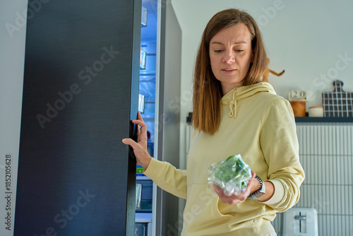 Woman standing by open refrigerator holding wrapped vegetables in modern kitchen