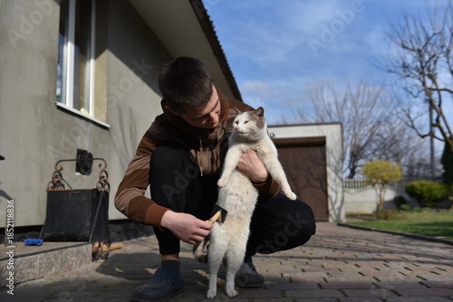 Young man brushing cat outdoors in yard, caring for pet near house on sunny day, calm domestic animal grooming moment