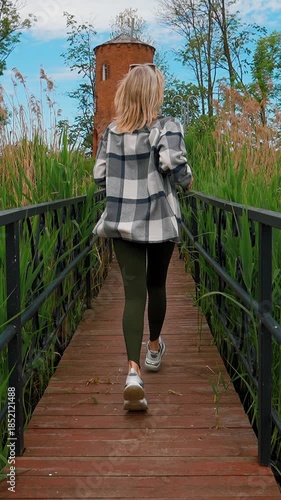 A young woman with blond hair runs casually across a wooden bridge and a stone path towards an old brick tower while enjoying a beautiful sunny day in a picturesque public park vertical video