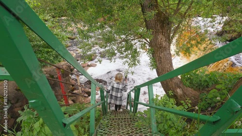 Young woman carefully descending a steep green metal staircase to a powerful flowing river in a lush green forest, exploring the natural landscape and capturing the moment on her phone belarus river v