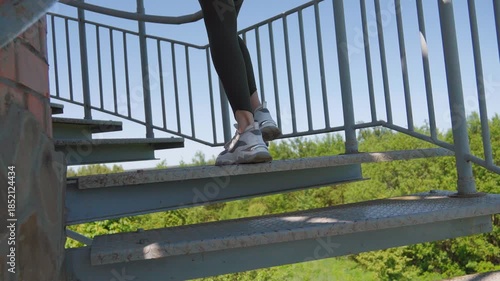 The feet of a young athletic woman in sneakers climb an outdoor metal spiral staircase, against the background of a green forest and a blue sky, showing the concept of a healthy and active lifestyle
