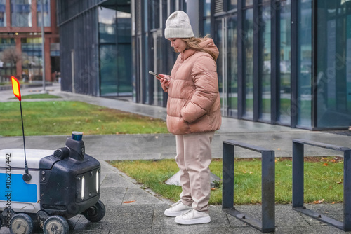 Smart robot courier stops for customer pickup. User reads smartphone message for delivery notification, tracking update. Contactless logistics transport moves through rainy street on wet pavement.