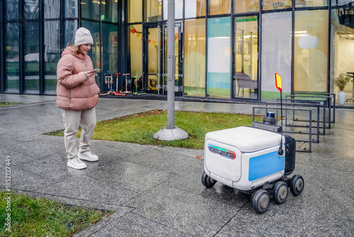 Autonomous delivery robot waits on sidewalk. Customer checks smartphone app for parcel status, qr verification. Modern urban courier service, contactless logistics transport on wet pavement.