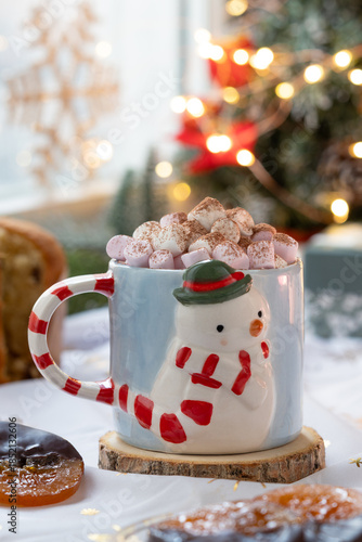 A mug of hot chocolate top with marshmallow and cacao powder with candied orange, cozy Christmas decoration atmosphere near window with light bokeh in background.