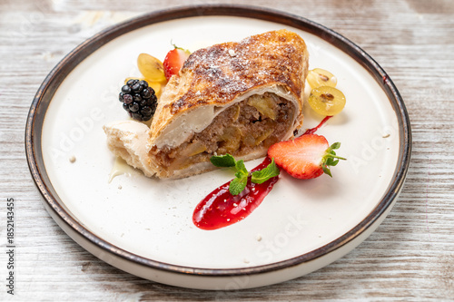 Slice of traditional apple strudel with flaky pastry and spiced apple filling, served on a ceramic plate with red berry sauce and fresh fruit. Elegant dessert still life, natural ligh