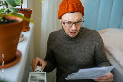 Fotografía Man in orange hat and warm sweater adjusting a radiator thermostat, looking shoc