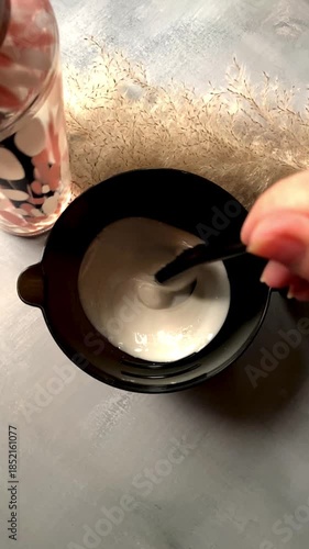 Close-up of hair dye mixture in a black bowl with professional coloring brush. Creamy beige hair color with soft light, dark background and dried pampas grass, creating a calm salon atmosphere.