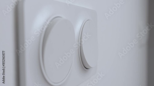 Close-up of hand pressing modern white wall light switch on interior wall to turn on indoor lighting. Concept of electricity use, home control and energy consumption