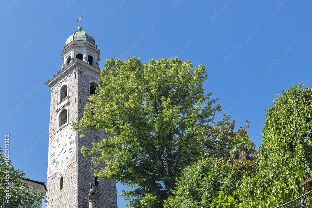 Obraz premium Stone Clock Tower With Green Dome And Cross Under Clear Blue Sky Surrounded By Trees, Lugano Cathedral, Cattedrale di San Lorenzo, Lugano, Switzerland