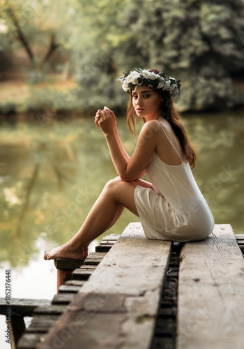 Bild auf Leinwand Beautiful girl in a white slip dress and floral crown sitting on a wooden jetty