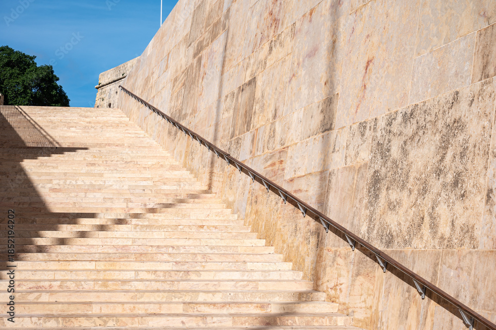 Fototapeta premium Historic ochre-colored stone stairs at the Parliament of Valletta, showcasing geometric patterns and shadows Valletta, Malta, 6 DEC 2025.