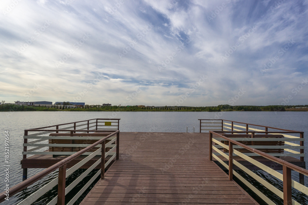 Fototapeta premium A wooden pier reaches out over still water. Trees and buildings line the far shore under a cloudy sky. The scene shows a quiet outdoor space for walking or fishing.
