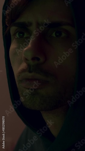 Introspective young man with dark hoodie gazes thoughtfully to the side, as camera pans smoothly, capturing emotional depth and atmosphere in a dimly lit room