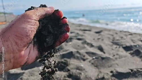 Person's hand is shown holding dark volcanic sand, letting it trickle through their fingers. In the background, the sparkling sea is visible, adding to the peaceful scene