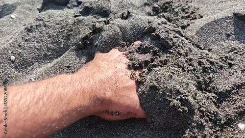 Close up of a man's hand slowly sinking and getting buried under the dark, coarse sand of a volcanic beach during a sunny summer day