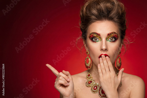 Glamorous woman surprised with colorful makeup and jewelry on red background  