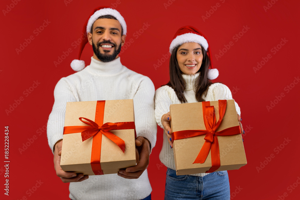 Fototapeta premium An arab couple, wearing Christmas hats, smiles joyfully while extending beautifully wrapped gifts towards the camera. The vibrant red background enhances their celebration of the winter holidays.
