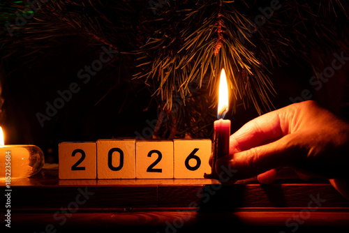 New Year tree and a woman's hand holding a candle near the tree and white cubes with the year 2026 in the dark