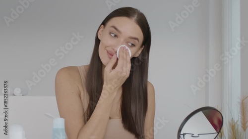 A woman with long hair gently uses a cotton pad to apply skincare in a bright, modern room. She sits in front of a mirror, surrounded by minimal decor, creating a calming atmosphere.