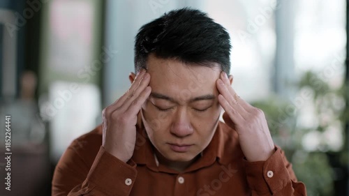 Inside a cozy cafe, a man sits with his eyes closed, holding his head with both hands. He appears to be feeling stressed or anxious amid a warm and inviting environment.
