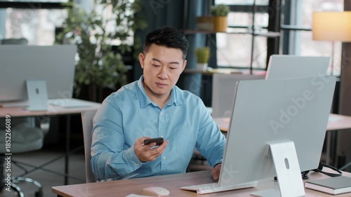 Bright office setting features a young man in a blue shirt using a computer and reaching for his smartphone. The space is modern and well-lit, with plants and a professional atmosphere.