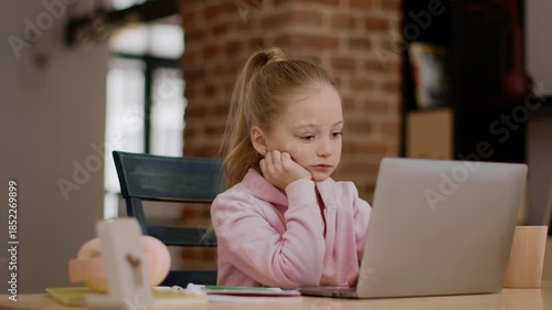 A young girl sits at a wooden table, resting her chin on her hand while looking at a laptop. She appears deep in thought, surrounded by school supplies in a bright room.