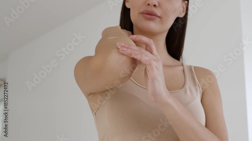 A woman in a light tank top tenderly applies lotion to her elbow while standing in a spacious and simple room with soft lighting. Her focus is on skincare and self-care.