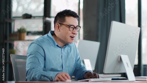 A focused man sits at a desk in a contemporary office, using two computers. Sunlight enters through large windows, creating a productive atmosphere for his tasks.
