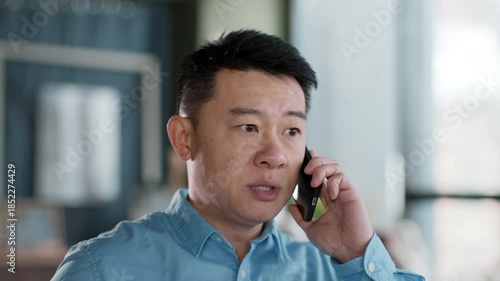 A businessman engaged in a phone conversation in a contemporary office setting. Natural light streams through large windows, highlighting the professional atmosphere.
