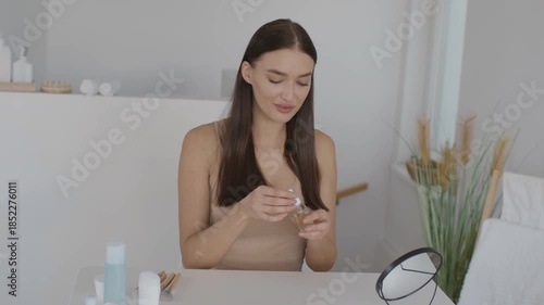 A woman is seated at a minimalist table in a well-lit room. She is applying serum from a small bottle, focused on her skincare routine. Natural light enhances the calming atmosphere.