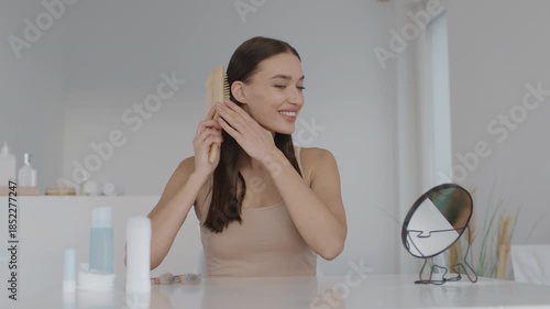 A woman is brushing her long hair at a clean, well-lit bathroom counter. She smiles while looking at her reflection in a round mirror. Beauty products are neatly arranged around her.
