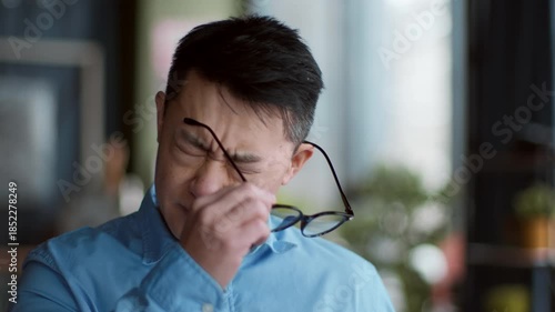A man in a blue shirt is squinting as he adjusts his glasses in a stylish cafe. The sunlight streams in through large windows, highlighting the relaxed atmosphere and greenery around him.