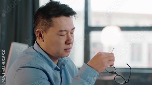 A man sits at a desk in a bright office, eyes closed in deep thought. He holds a pair of glasses in his hand. Natural light beams through large windows, creating a calm atmosphere.