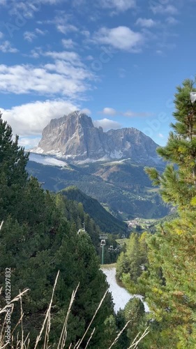Cable car moving through alpine landscape during summer season in the Dolomites, Italy. Scenic mountain view with green forests, valleys, rocky peaks, and clear blue sky