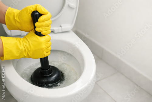 Clogged toilet being cleaned with rubber plunger and protective yellow gloves in household bathroom on white ceramic floor tiles