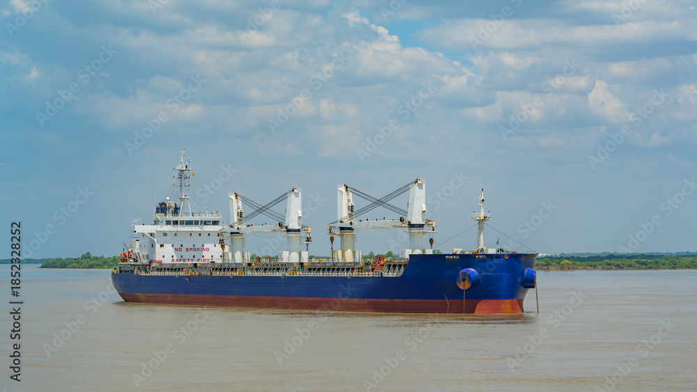 Naklejka premium Blue bulk carrier ship with four deck cranes anchored on a calm river under a cloudy sky. Industrial maritime transport and logistics photography