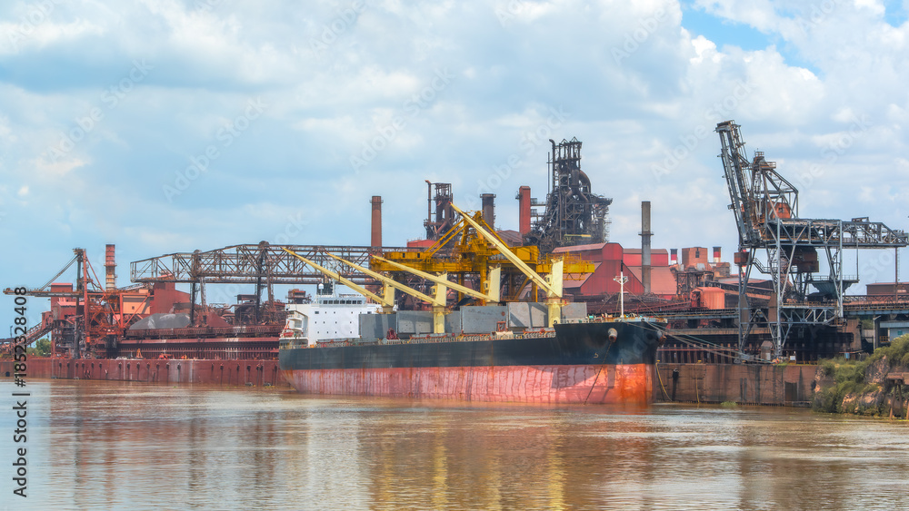 Fototapeta premium Bulk carrier ship docked at a large industrial port with a red steel mill and tall smokestacks under a bright, cloudy sky, reflecting in the brown river water