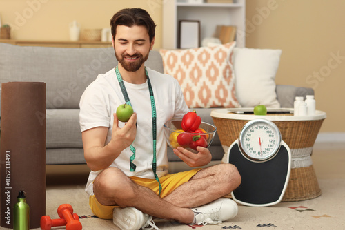 Sporty young man with healthy food and weight scales sitting at home