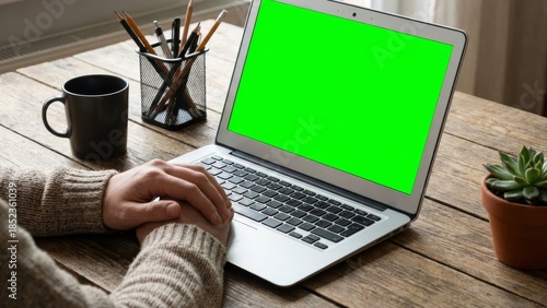 Person working on laptop with green screen on wooden table with office supplies