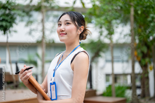 Free Download Asian woman office worker wearings lanyard holding pen and book while standing or walking in a park. - GFXLoad
