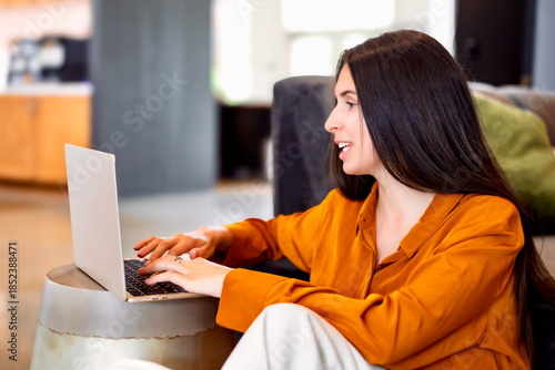 Smiling woman working on a laptop in a bright home office, sitting on the floor and enjoying a relaxed remote work moment with casual digital activity and a possible video call.