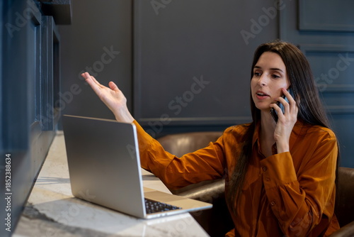 Frustrated woman having an argument during a phone call while working on a laptop in a modern workspace, expressing confusion and communication issues in a remote work environment.
