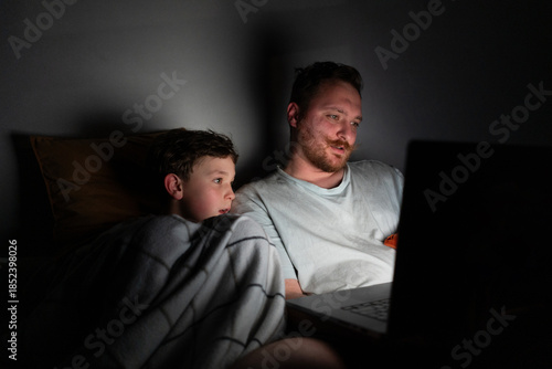A father and son sit closely on a bed, looking at a laptop screen. The room is dark, with soft light from the laptop illuminating their faces. They are sharing a moment of laughter and joy.