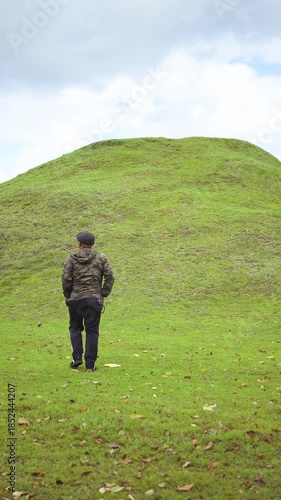 A male adventurer enjoys some alone time in a green grassy area with a small hill in front of him. An Asian male traveler daydreams alone in the peaceful and tranquil atmosphere of a green meadow.