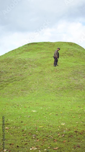 A male adventurer enjoys some alone time in a green grassy area with a small hill in front of him. An Asian male traveler daydreams alone in the peaceful and tranquil atmosphere of a green meadow.